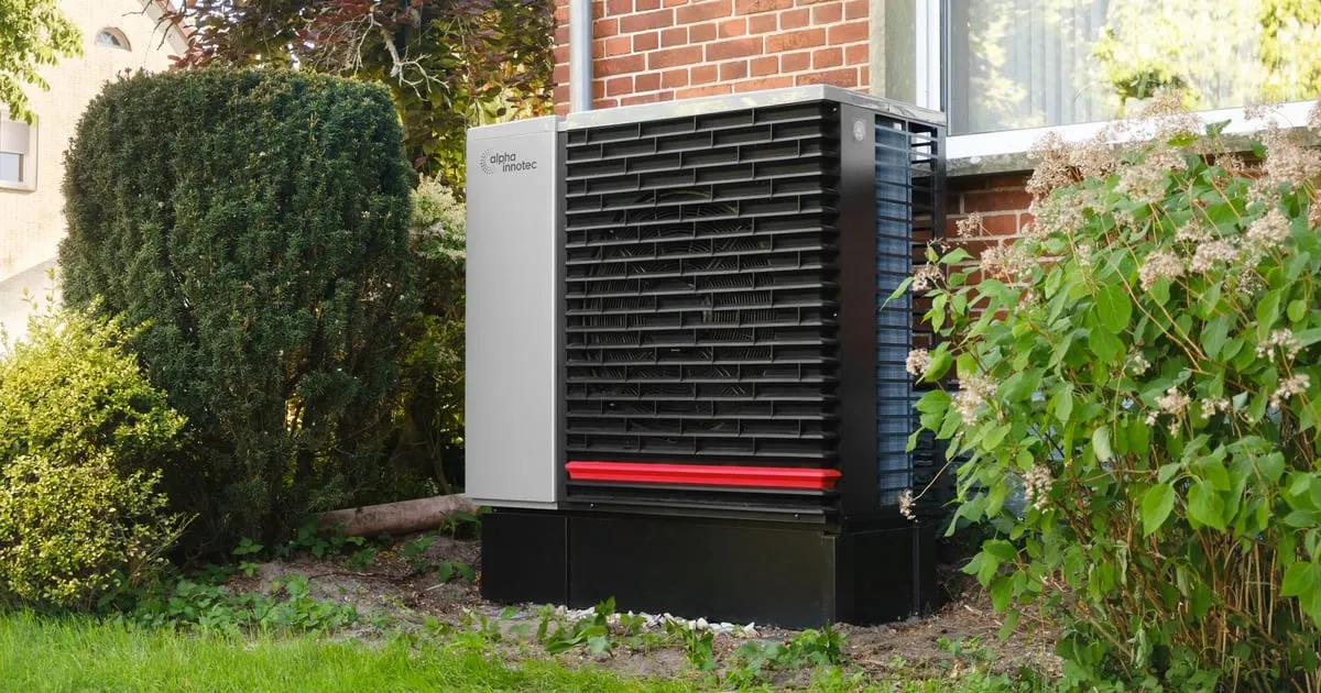 A domestic air source heat pump unit standing on a plinth in a garden beside a brick house
