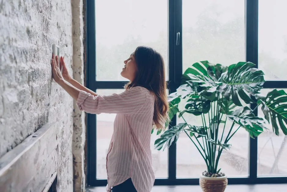 A woman adjusting a wall-mounted thermostat in a bright room with large windows
